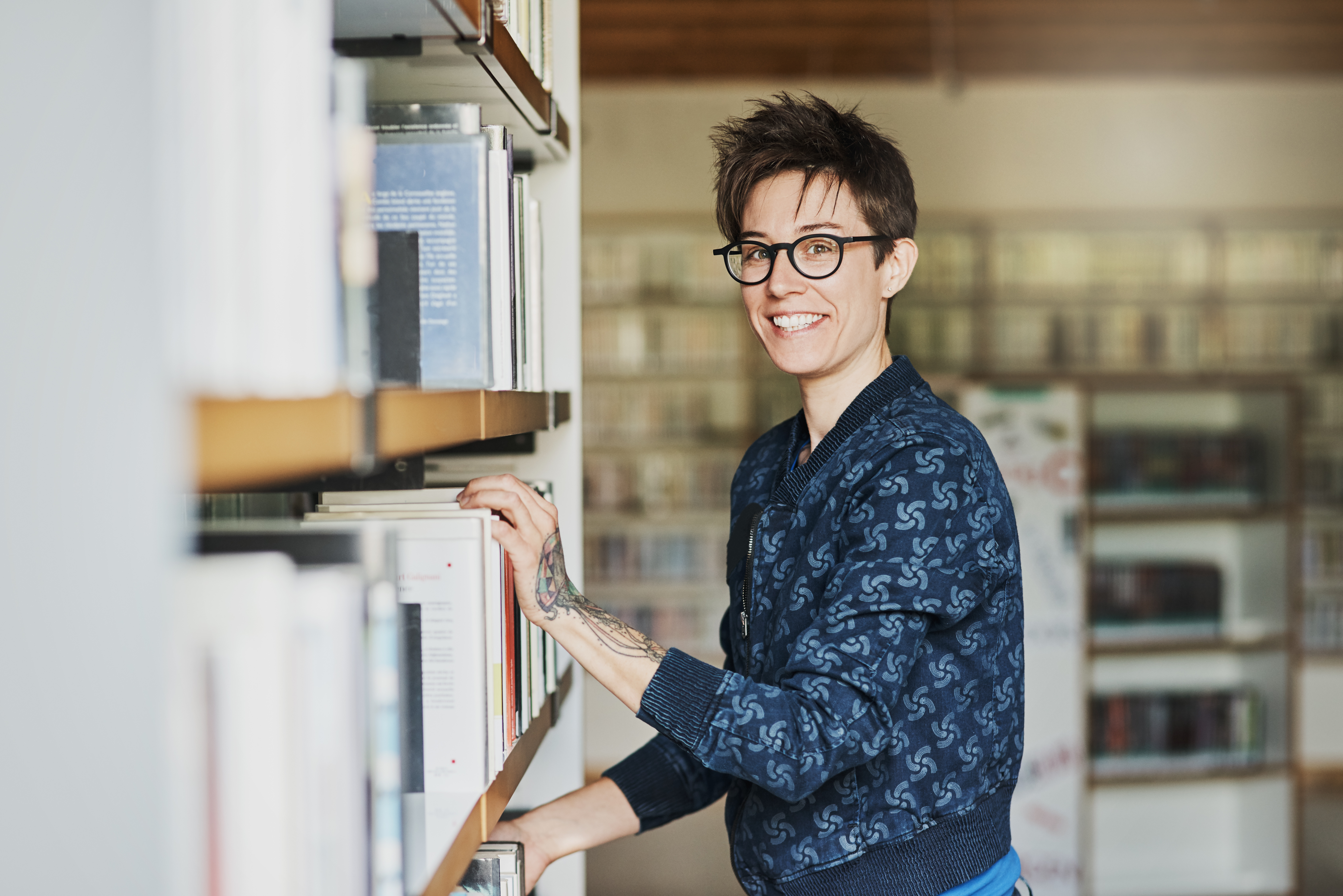 Person with glasses, standing and smiling next to library shelf.