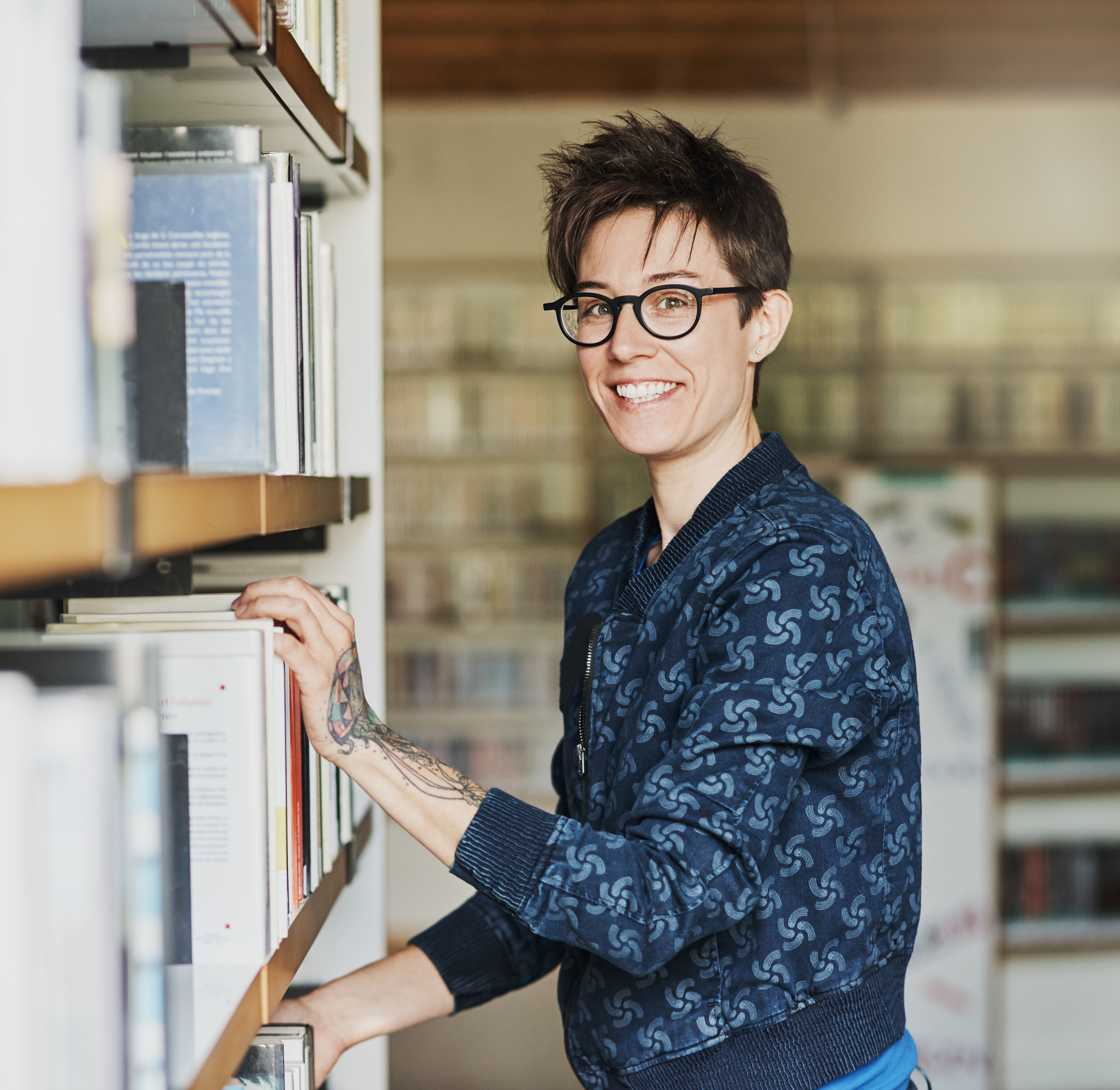 Person standing and smiling whilst browsing books at a library.